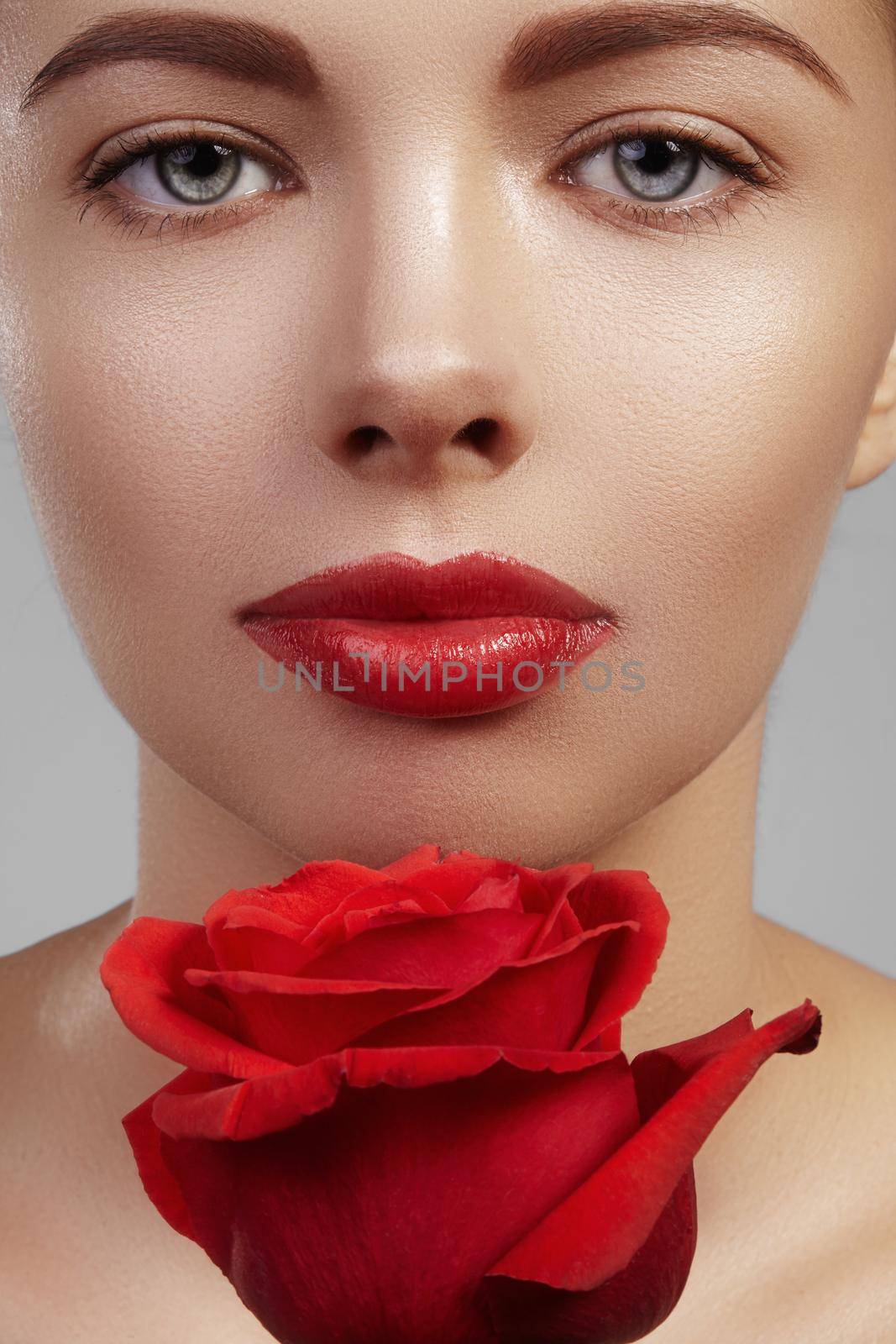 Close-up beautiful young woman with bright lipgloss makeup. Perfect clean skin, sexy red lip make-up. Beautiful valentine visage with red rose flower. Romantic and sexy look for Valentines day.