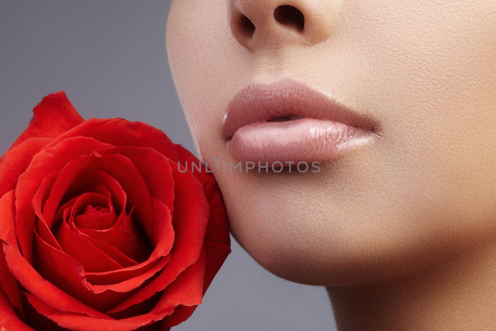 Close-up beautiful female lips with bright lipgloss makeup. Perfect clean skin, light fresh lip make-up. Beautiful spa portrait with red rose flower. Spa and cosmetics