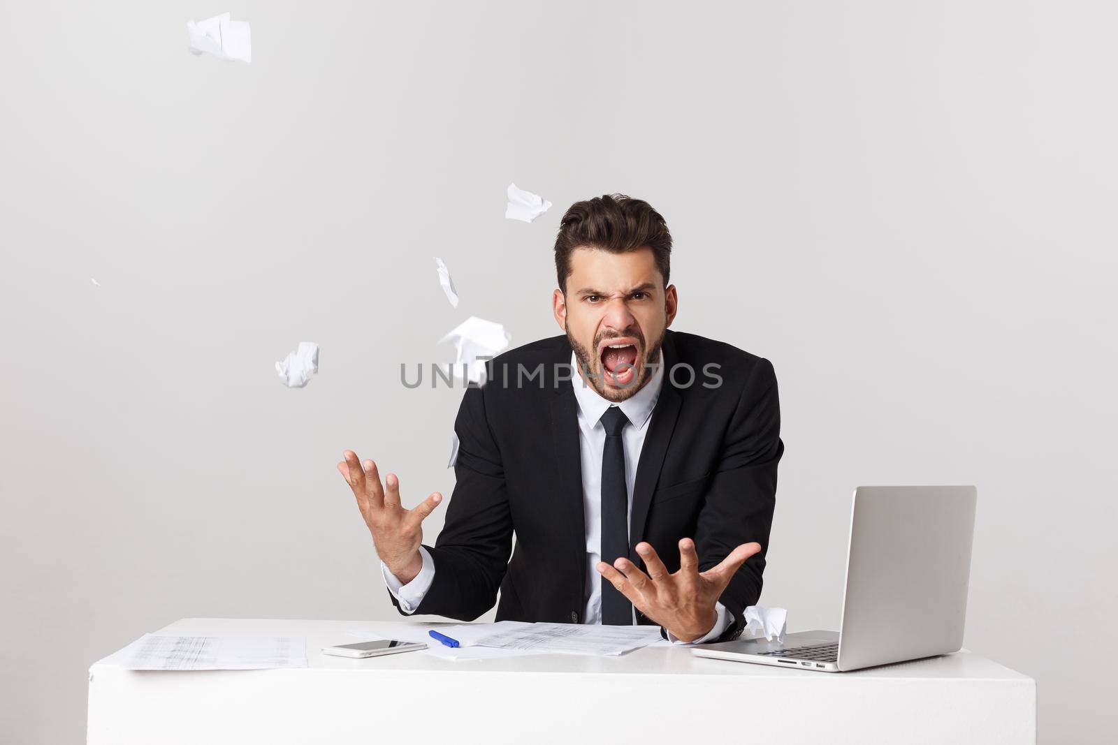 Angry young bearded man work at desk with laptop isolated over white background. Screaming tearing paper documents.