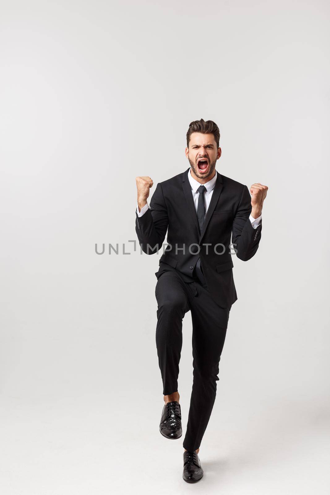 Cheerful young bearded business man show hand up excited with clenched fists. Full length portrait business man isolated over white studio background.