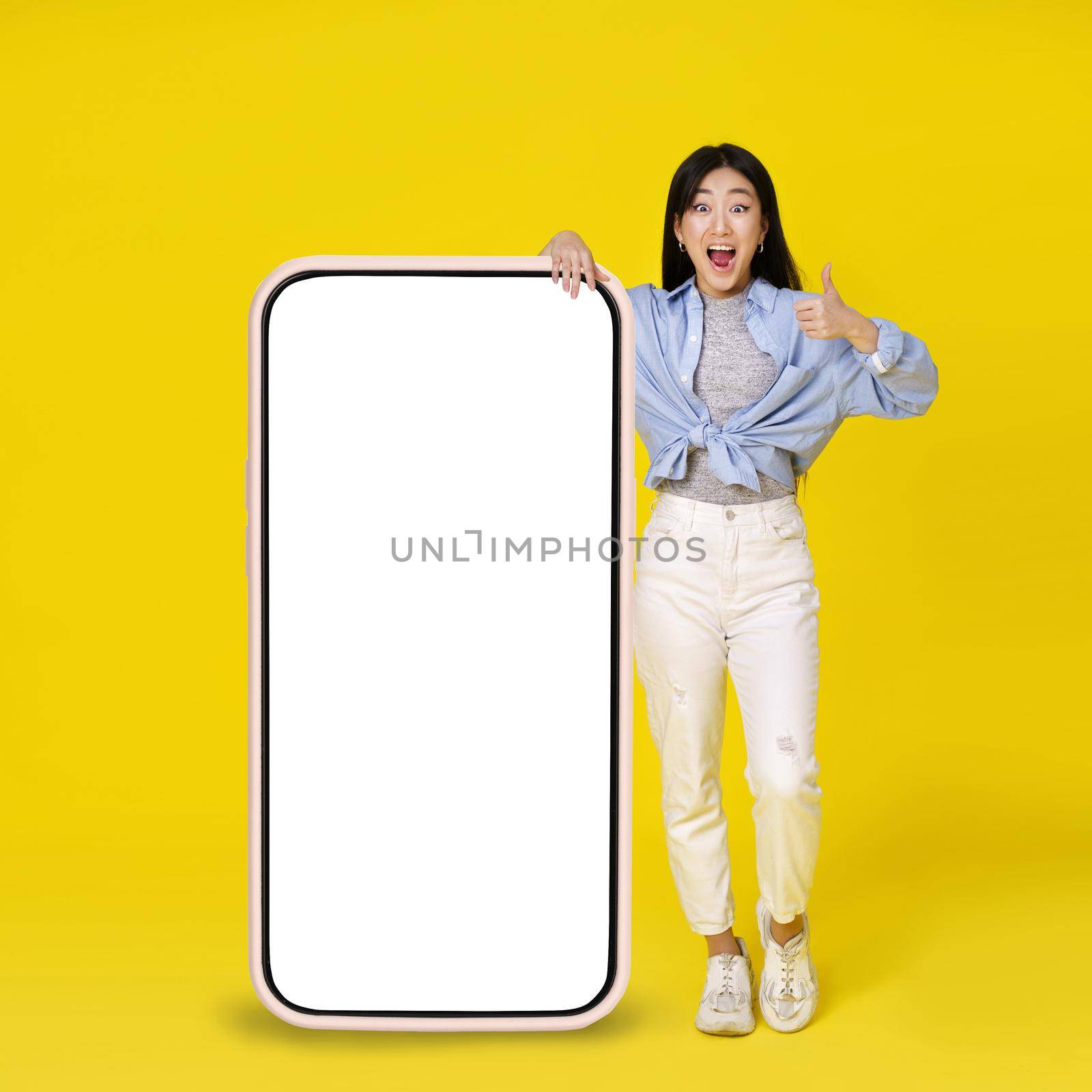 Excited asian girl gesturing thumb up leaned on giant, huge smartphone with white, blank screen standing leaned on wearing casual isolated on yellow background. Free space mock up by LipikStockMedia