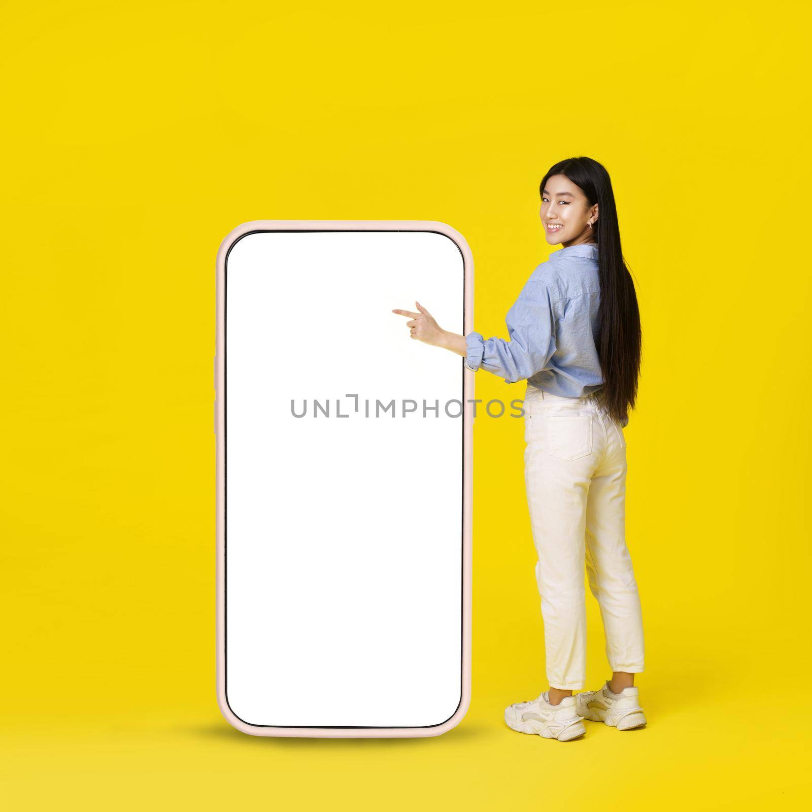 Asian girl pointing on giant, huge smartphone with white blank screen standing back happy pointing finger up dressed in casual isolated on yellow background. Free space mock up.