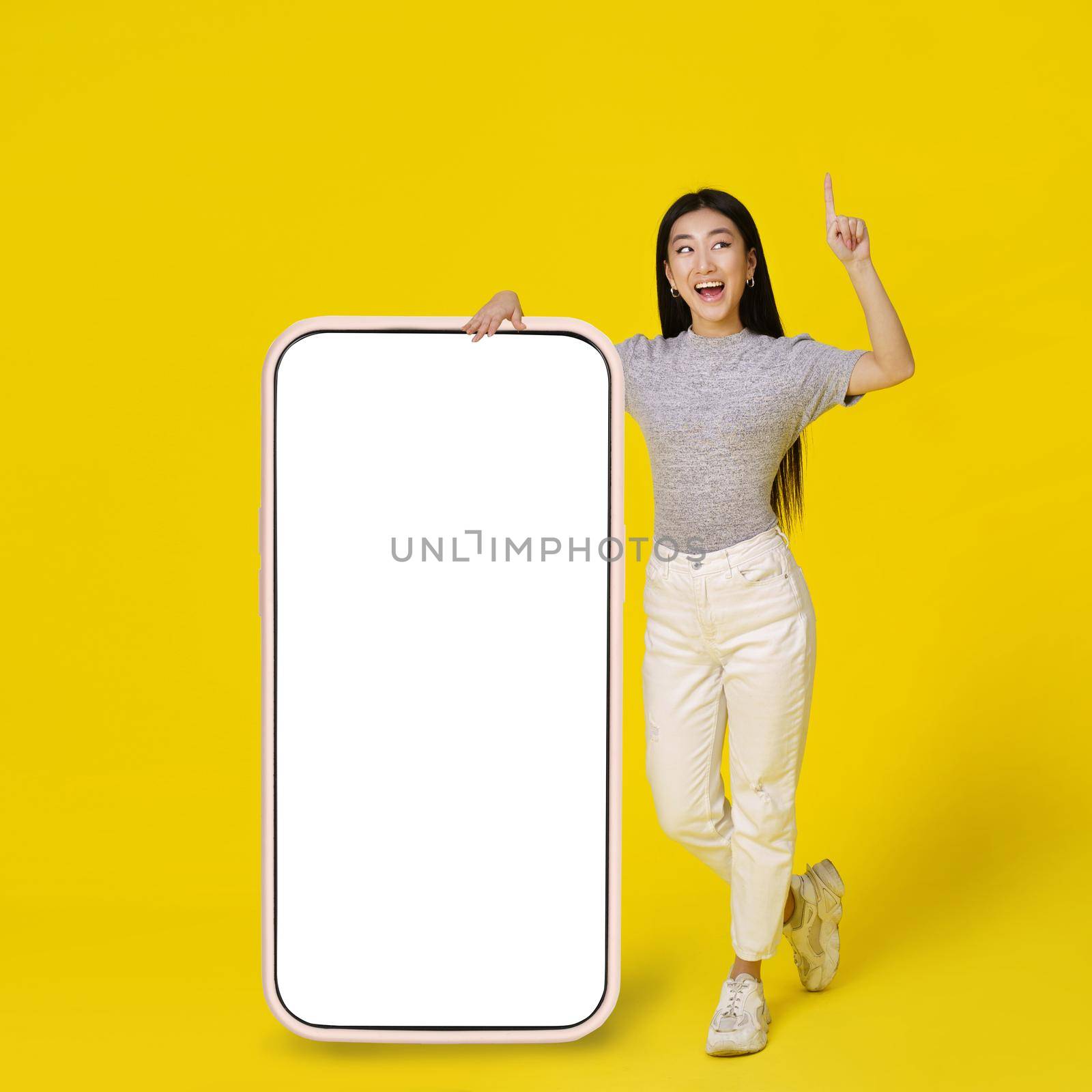 Happy asian girl leaned on giant, huge smartphone with white, blank screen happy pointing finger up dressed in casual isolated on yellow background. Free space mock up by LipikStockMedia