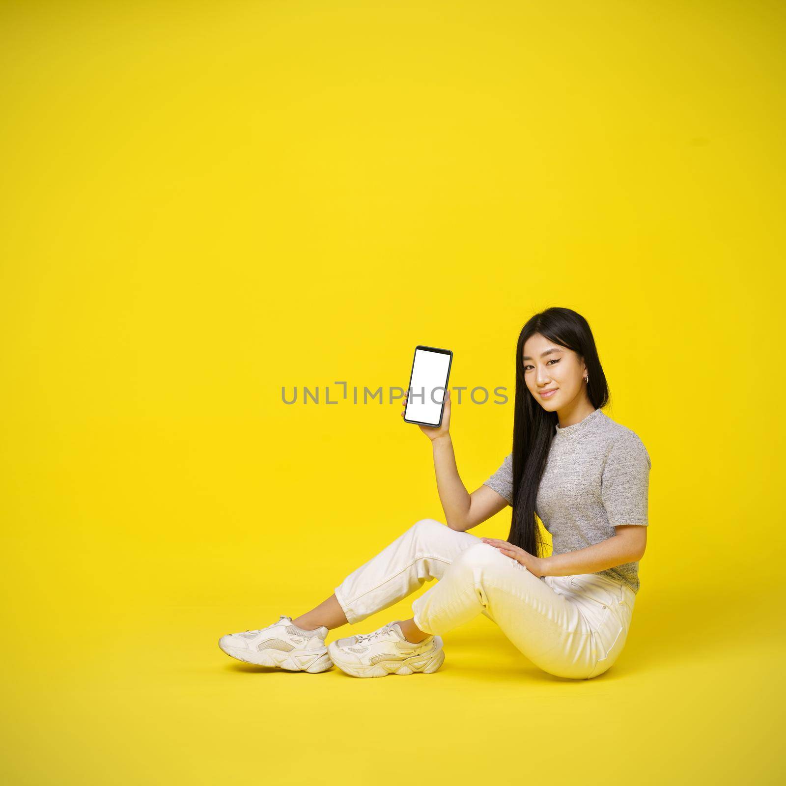 Charming asian young girl holding smartphone sitting on the floor showing a white screen isolated on yellow background. Great offer. Product placement. Mobile app advertisement. Copy space.