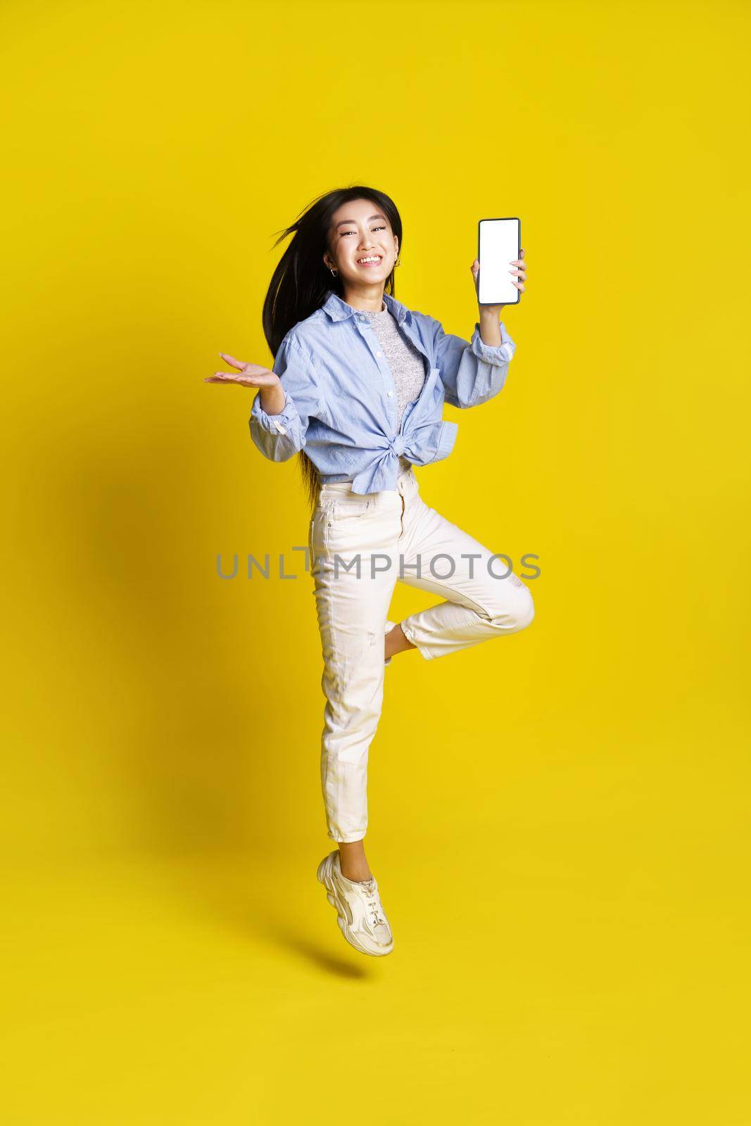 Happy asian girl jumping holding smartphone showing white screen mobile app advertisement isolated on yellow background. Full length portrait of joyful asian girl. Product placement.