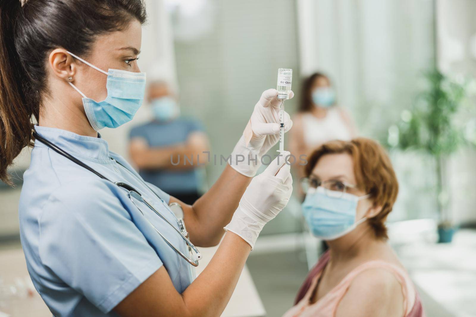 Nurse holding syringe and making Covid-19 vaccination injection dose in shoulder of female senior patient wearing mask.