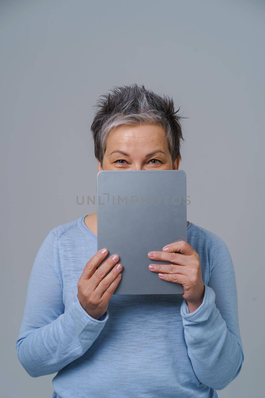 Charming mature grey haired woman hide shy behind digital tablet working or shopping online or checking on social media. Pretty woman in blue blouse isolated on white background.