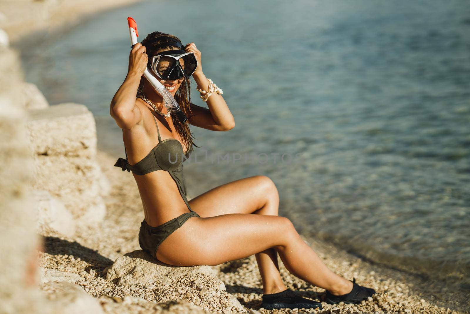 An attractive young woman sitting by the water with her scuba mask and snorkel and preparing to snorkeling in the sea.