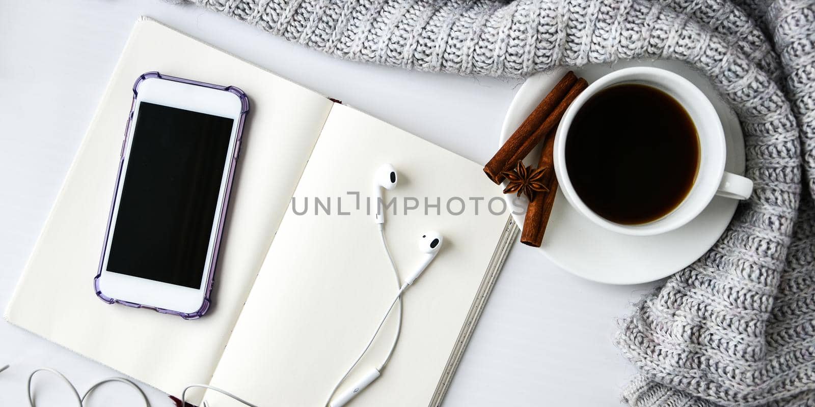 Cup of coffee with notebook phone headphones cinnamon sticks and anise star on white background. Sweater around. Winter morning routine. Coffee break. Copy space. Top view. Breakfast. Flat lay