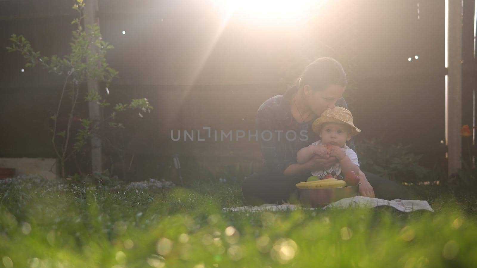 Happy Young Cheerful Mother Holding Baby Eating Fruits On Green Grass. Mom Adorable Infant Child Playing Outdoors With Love In Backyard Garden. Little Kid With Parents. Family, Nature, Ecology Concept by mytrykau