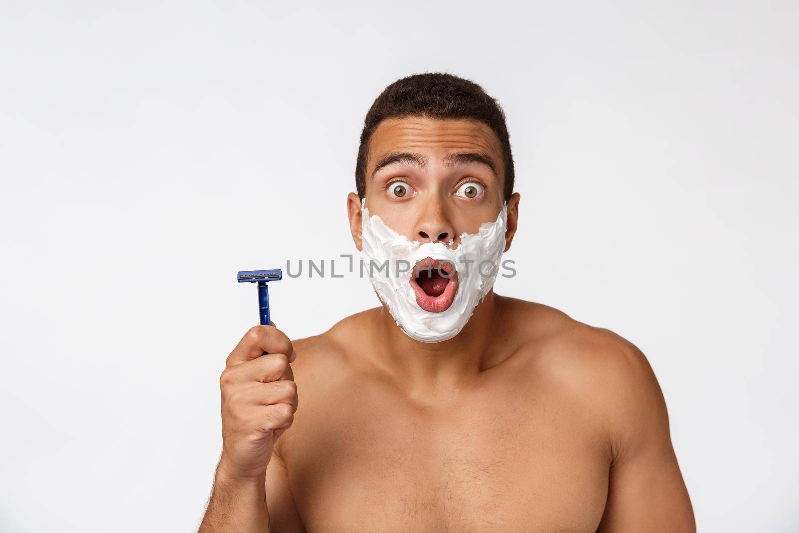Close up of a happy naked african man with face in shaving foam holding razor isolated over gray background. by Benzoix