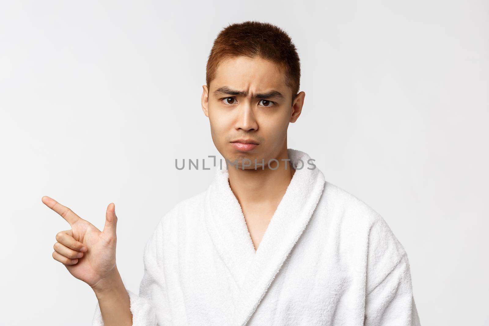 Beauty, spa and leisure concept. Portrait of skeptical, upset and moody asian man in bathrobe, sighing and frowning, sulking disappointed, pointing finger left at something bad, white background.