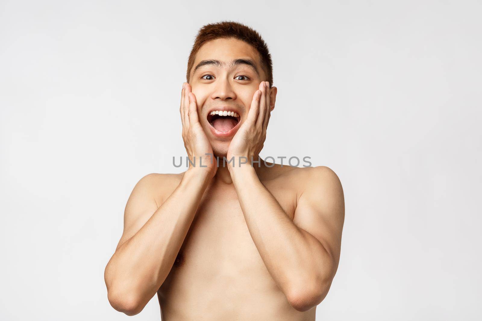 Beauty, people and home concept. Close-up portrait of amused naked asian guy, smiling excited, open mouth fascinated, touch cheeks and staring camera amazed, standing white background by Benzoix