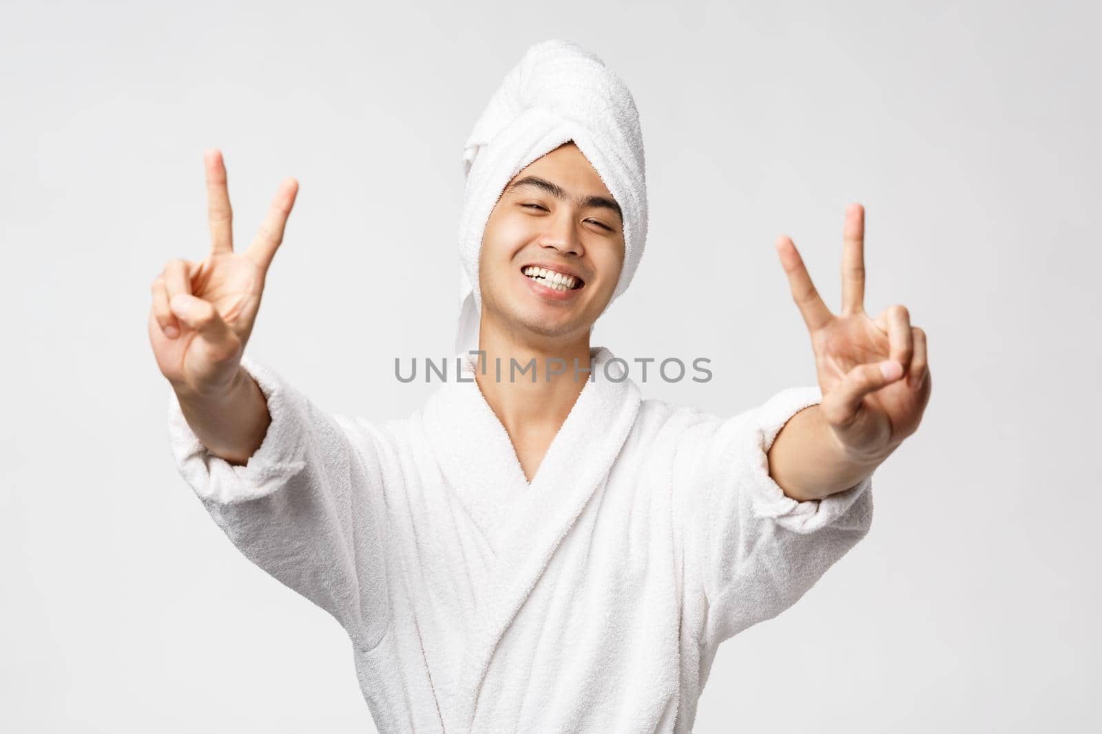 Beauty, spa and leisure concept. Upbeat handsome asian man in bathrobe and bath towel, show peace signs and smiling with rejoice, express enthusiasm, stand white background.