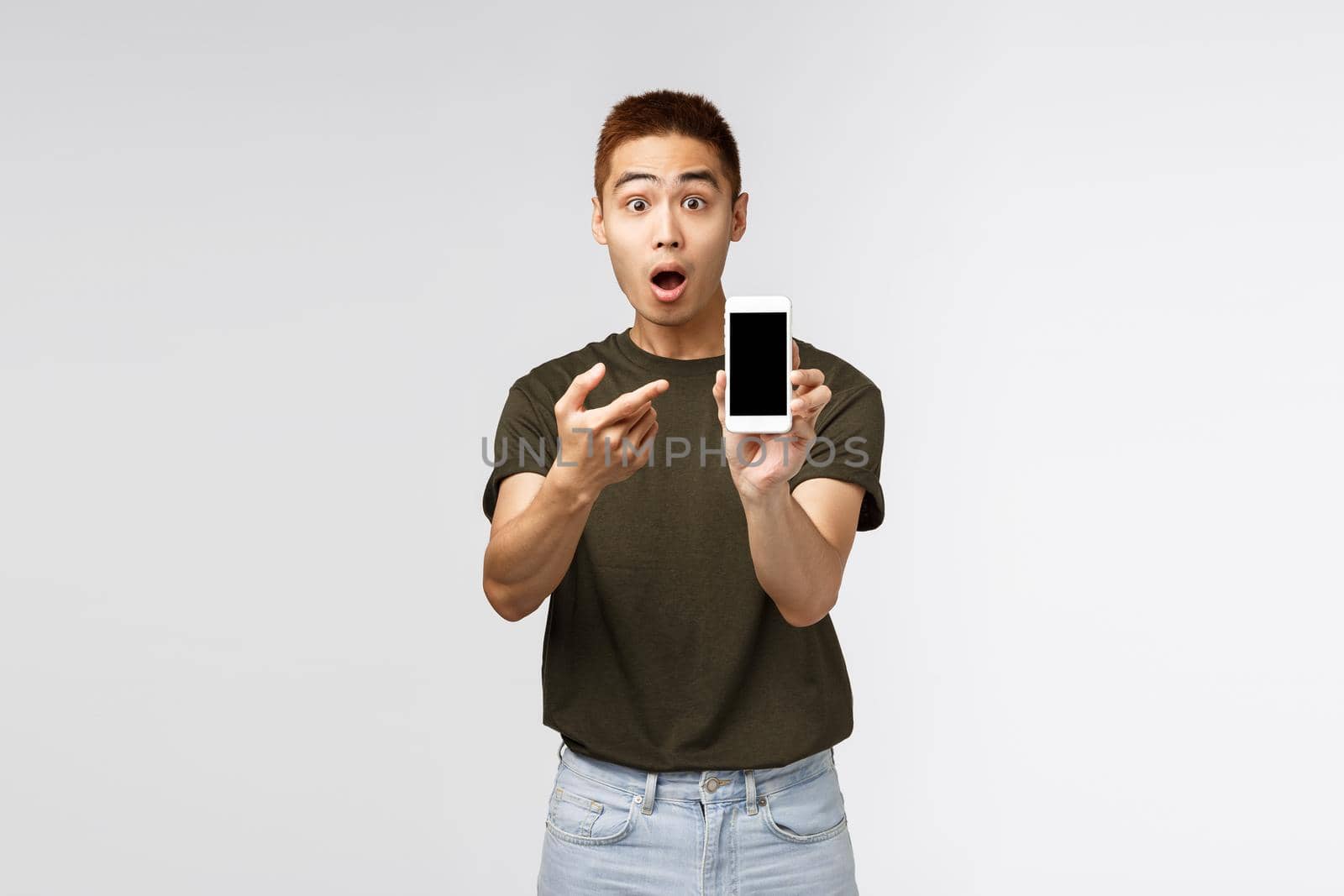 Technology, online lifestyle and communication concept. Surprised and amazed asian guy shocked pointing at mobile phone display to show something stunning and amazing, grey background by Benzoix