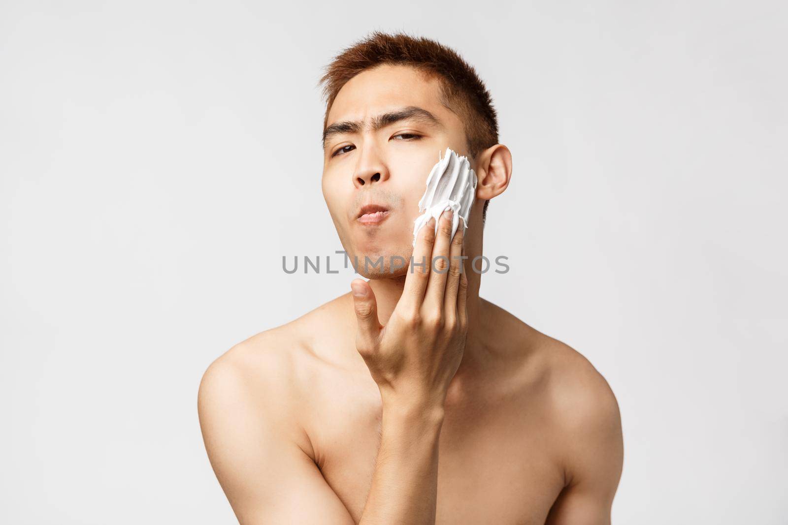 Beauty, people and hygiene concept. Portrait of funny handsome young man with naked torso, looking at bathroom mirror, apply shaving cream to face, standing white background.