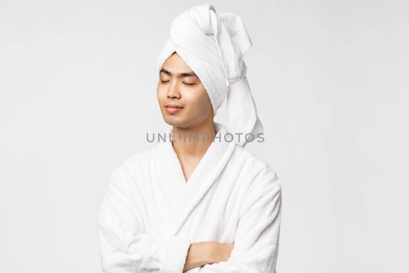 Beauty, spa and leisure concept. Portrait of relaxed, calm and peaceful happy asian man enjoying vacation, close eyes and smiling, wear bathrobe and towel over head, white background by Benzoix