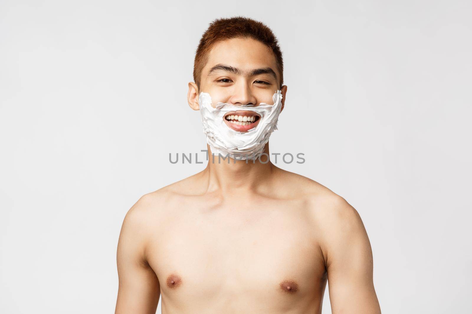 Beauty, people and hygiene concept. Portrait of handsome pleased asian man with naked torso, getting ready to work, smiling happy with shaving cream on face, standing white background.