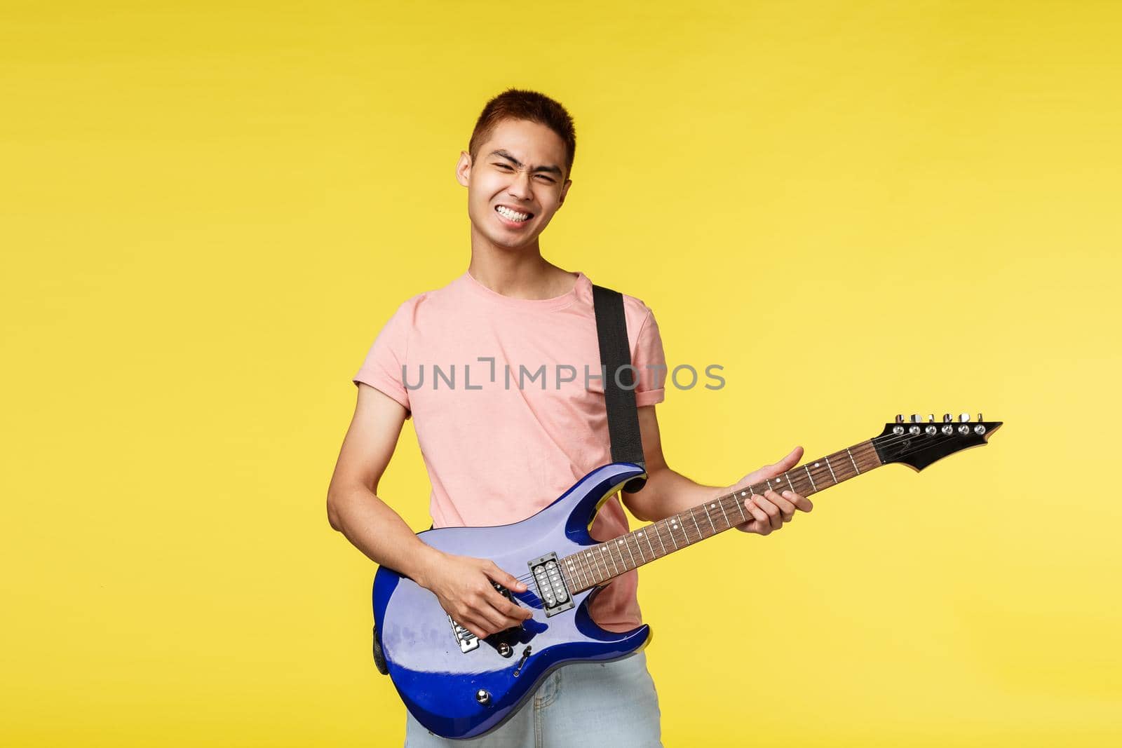 Lifestyle, leisure and youth concept. Lets jam. Carefree smiling asian guy playing in band, holding blue electric guitar, feel rock-n-roll start on stage, standing upbeat yellow background.