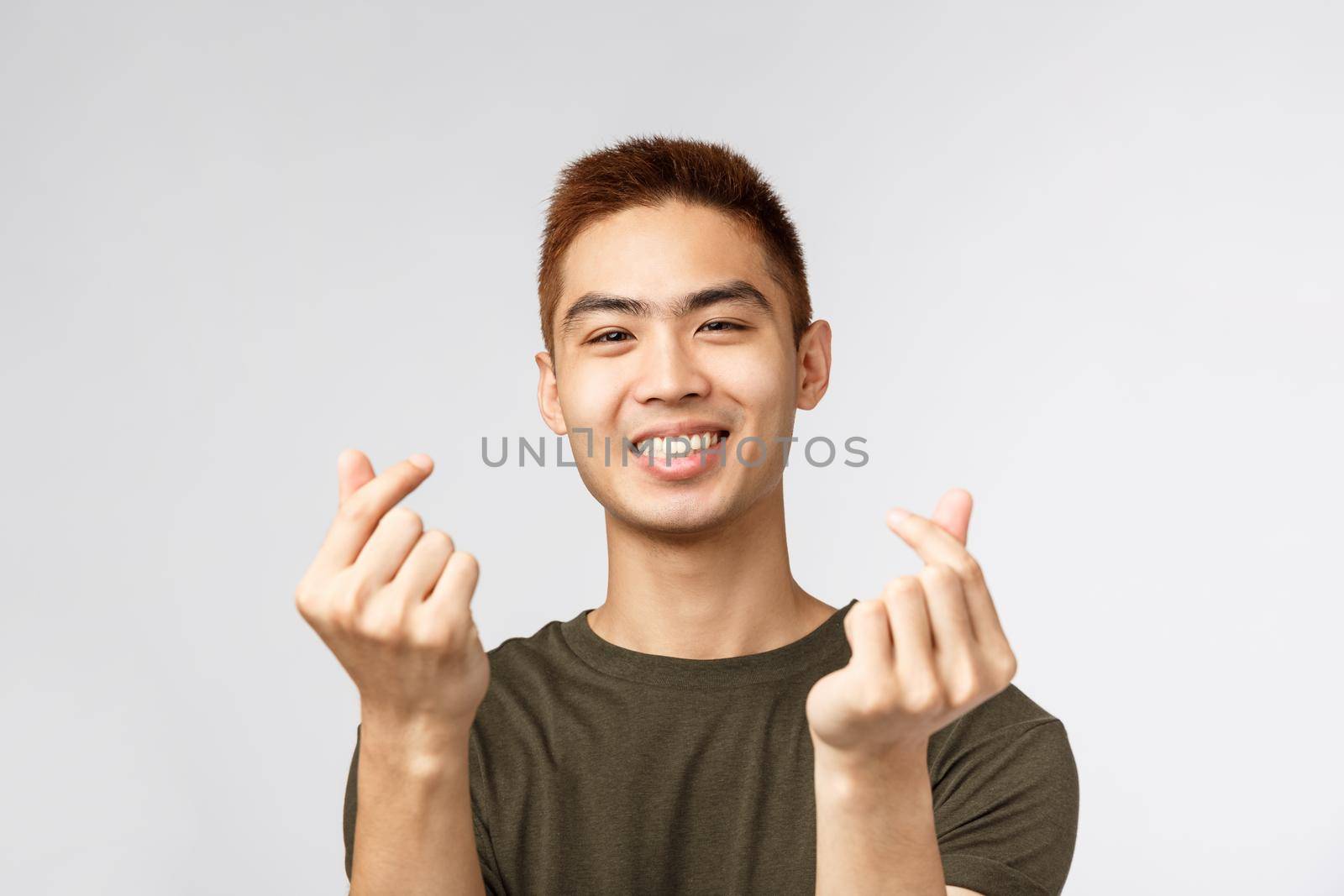 People, different expressions and lifestyle concept. Cute asian guy with stylish haircut, showing korean finger hearts and smiling lovely, express sympathy or like, stand grey background.