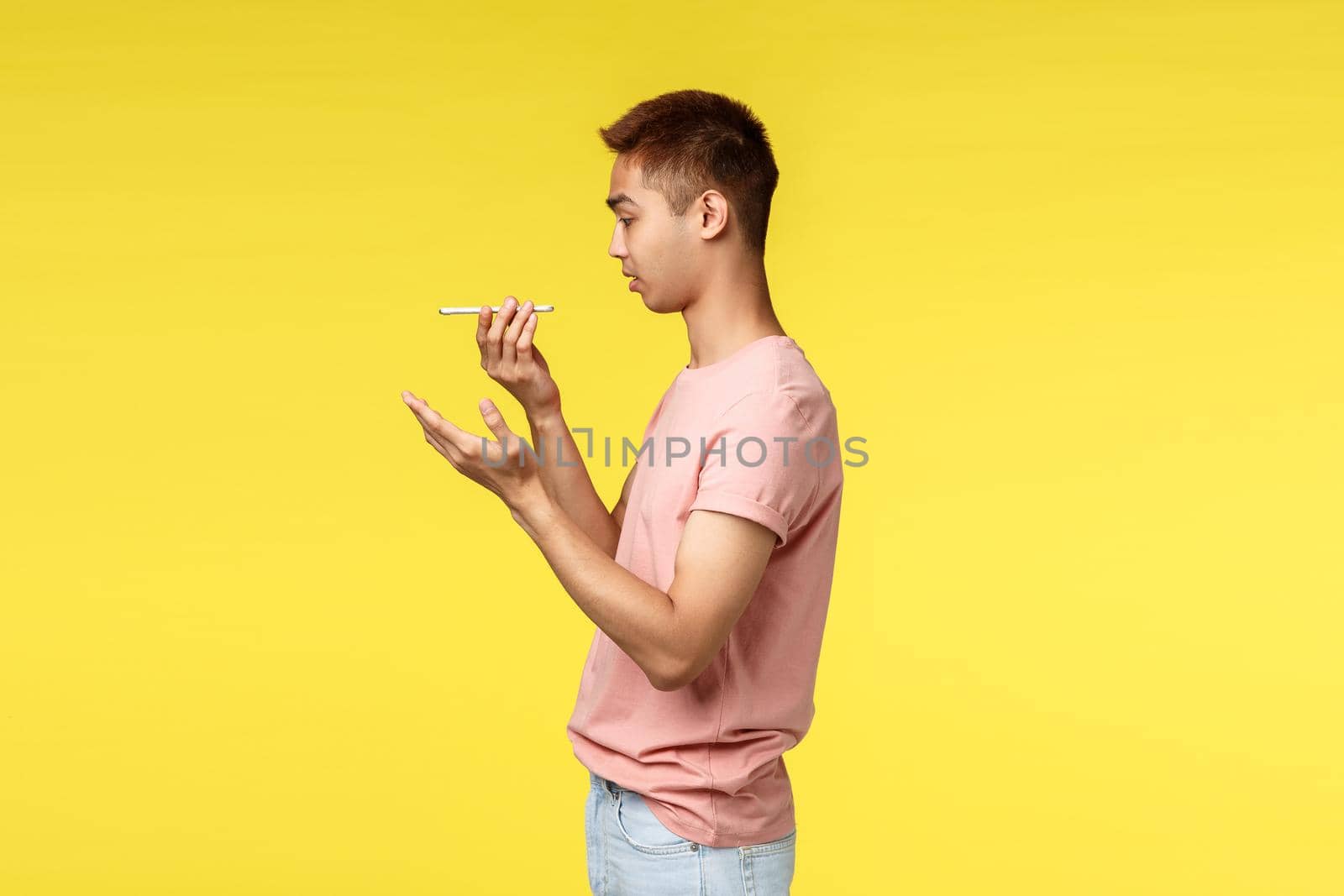Technology, communication and lifestyle concept. Profile portrait of asian hipster guy in pink t-shirt, using mobile phone, leaving voice message, speaking into speaker smartphone, yellow background.