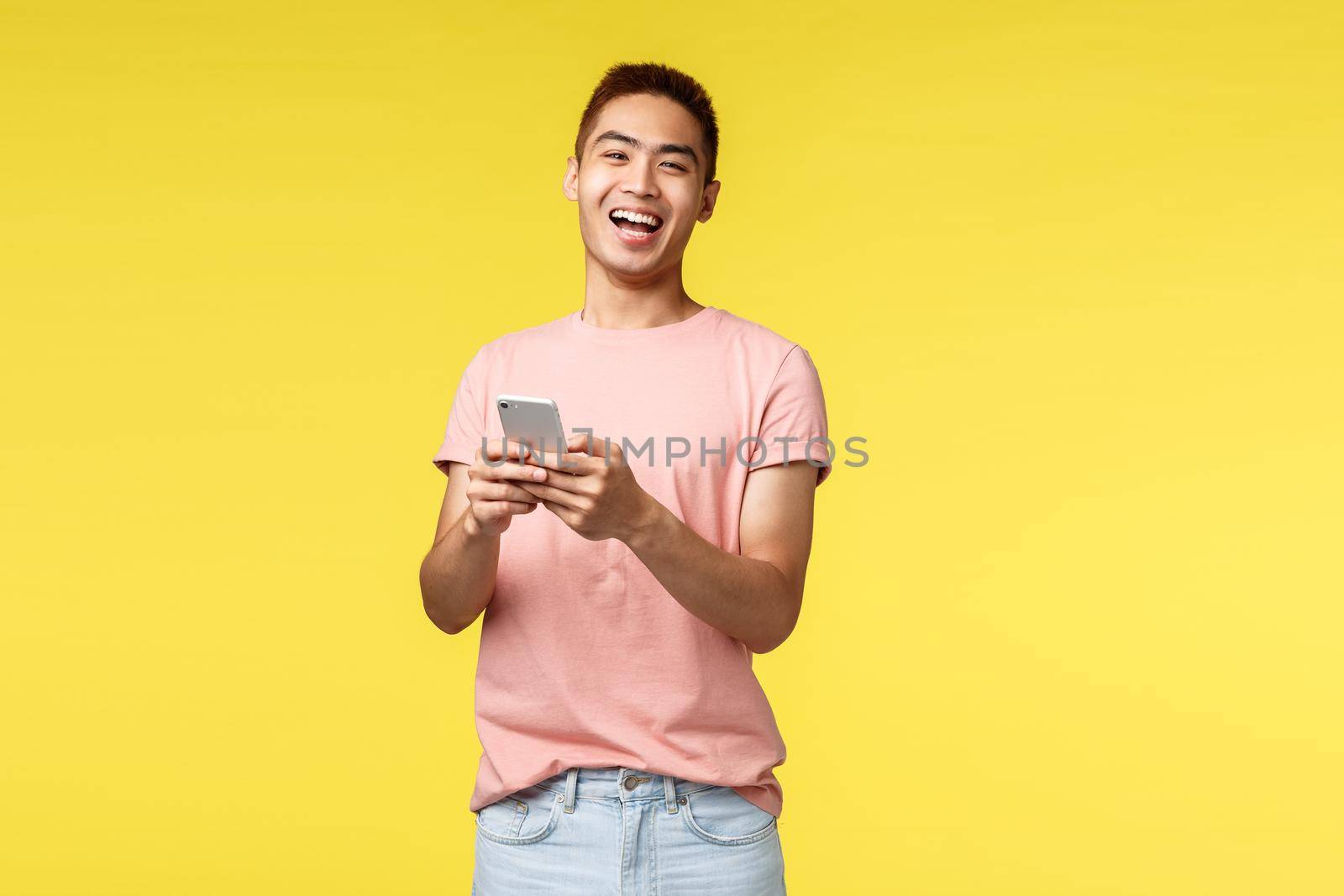 Technology, communication and lifestyle concept. Enthusiastic asian male in pink t-shirt laughing from cool joke or video, using mobile phone, smiling pleased camera, stand yellow background.