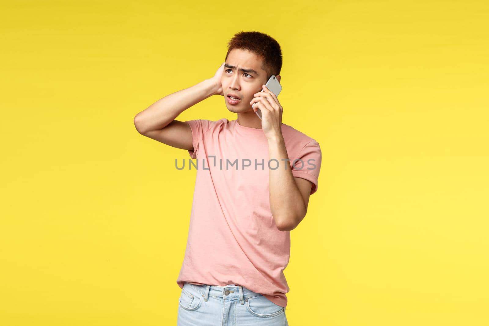 Technology, communication and lifestyle concept. Portrait of troubled, indecisive young asian guy look puzzled up, talking on mobile phone, having tough conversation, yellow background by Benzoix
