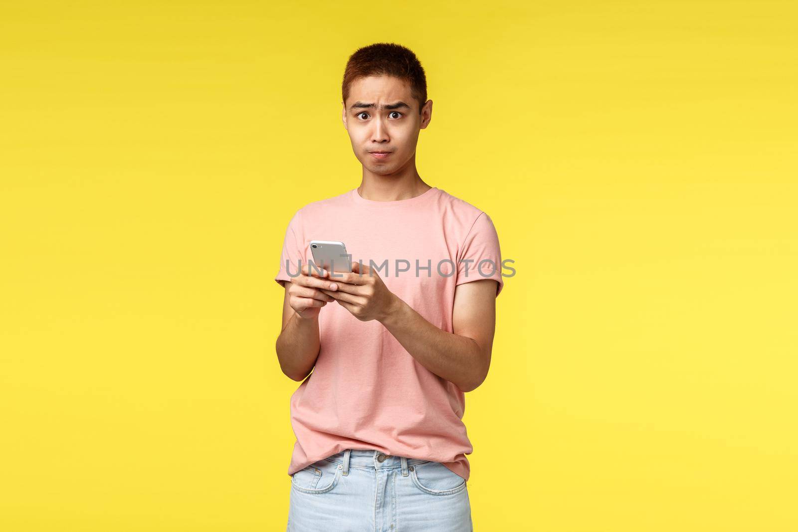 Technology, communication and lifestyle concept. Portrait of alarmed asian guy in pink t-shirt, using mobile phone text alarmed at bad news he read internet, standing bothered and frustrated.
