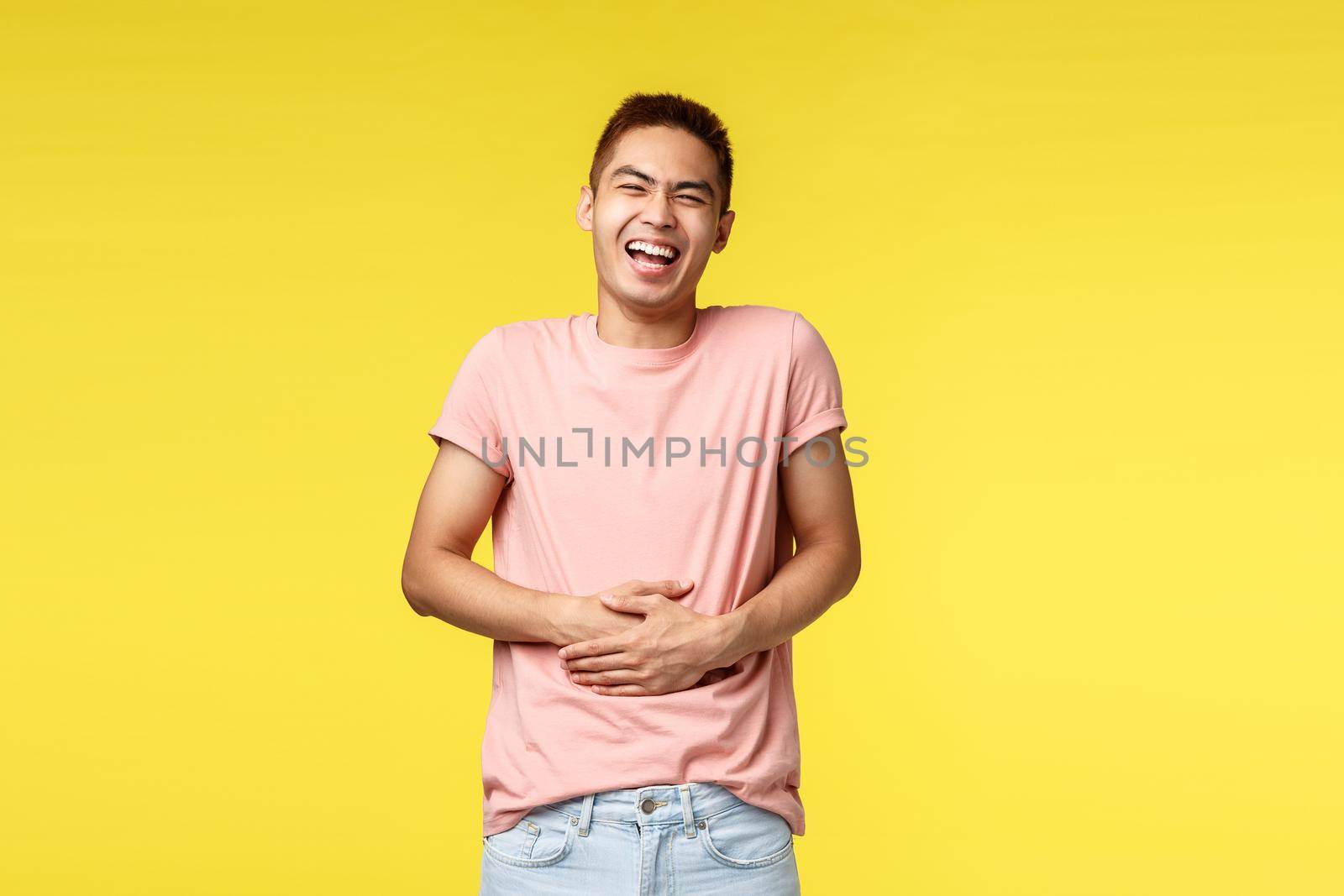 Lifestyle, travel and people concept. lmao so funny. Portrait of cheerful, enthusiastic asian man in pink t-shirt, laughing out loud, belly aches from giggle, smiling broadly, yellow background.