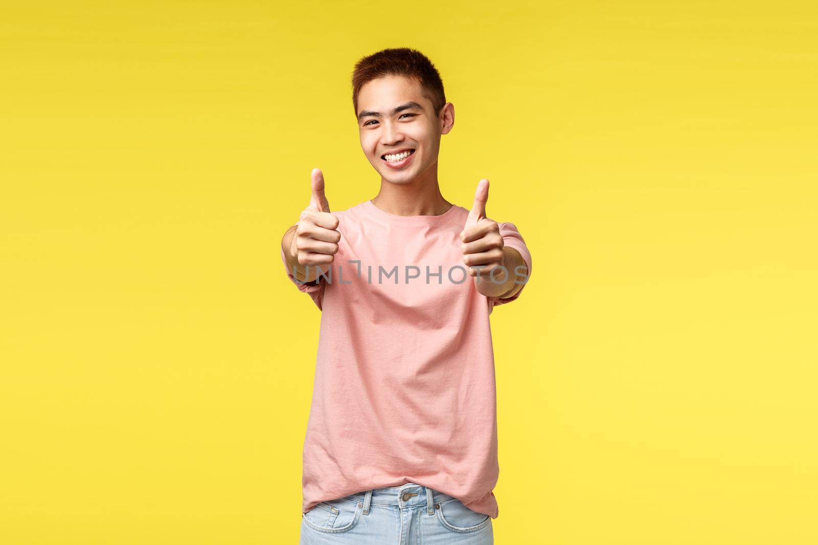 Lifestyle, travel and people concept. Satisfied cute asian male student recommend best language courses in town, show guarantee or approval sign, thumb-up smiling pleased, yellow background.