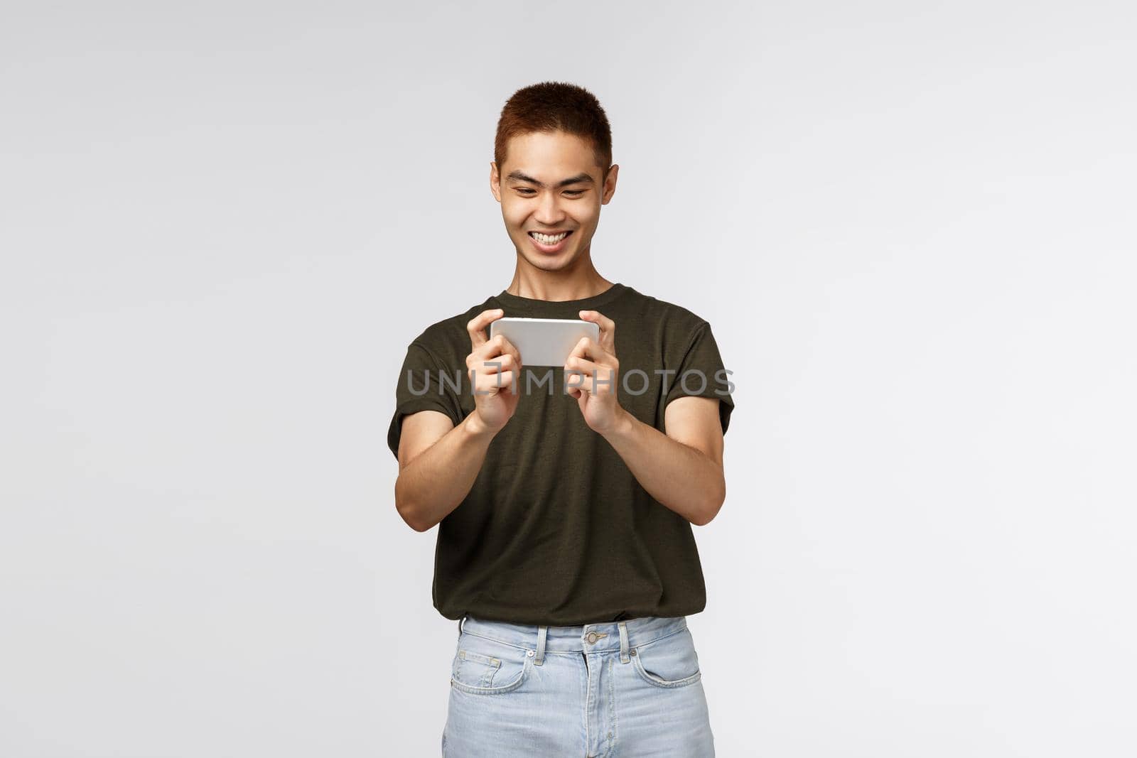 Technology, online lifestyle and communication concept. Happy pleased young asian male using mobile phone, smiling cheerful as playing new smartphone app, grin satisfied, grey background.