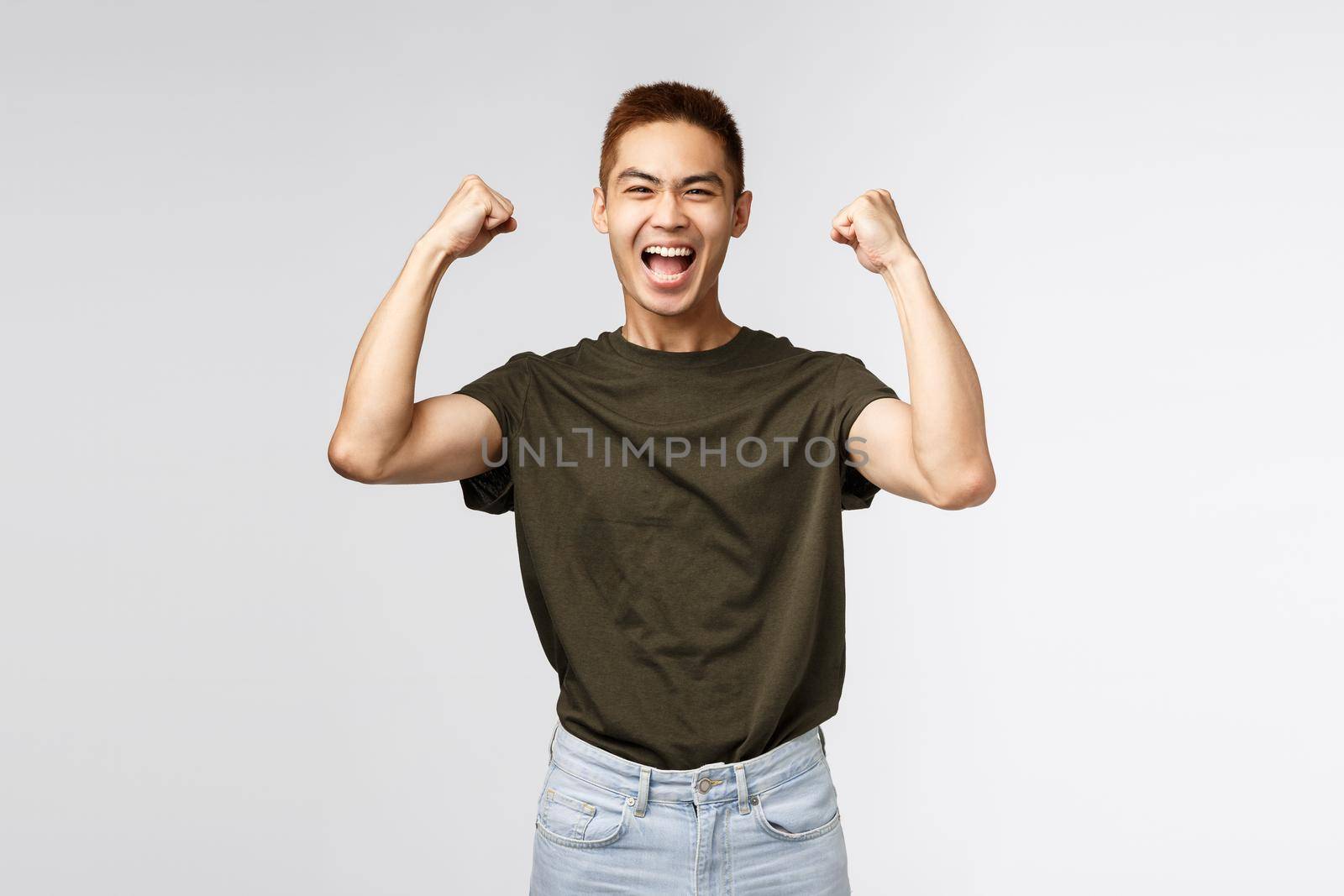 Competition, prize and people concept. Portrait of happy, triumphing young asian man raising hands up, fist pump celebrating victory, achieve success and winning, smiling upbeat, grey background by Benzoix