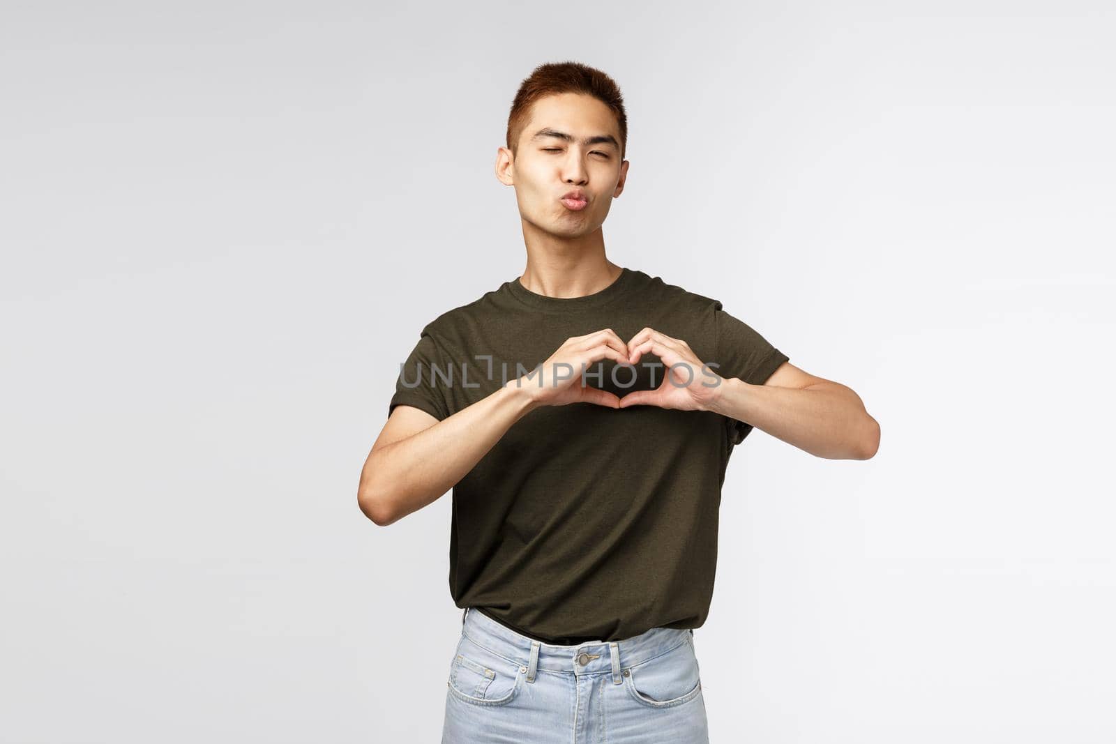 People, emotions and lifestyle concept. Cute and silly male student, asian guy wink cheeky and flirty, pouting or folding lips for kiss and showing heart over chest, express sympathy and love.
