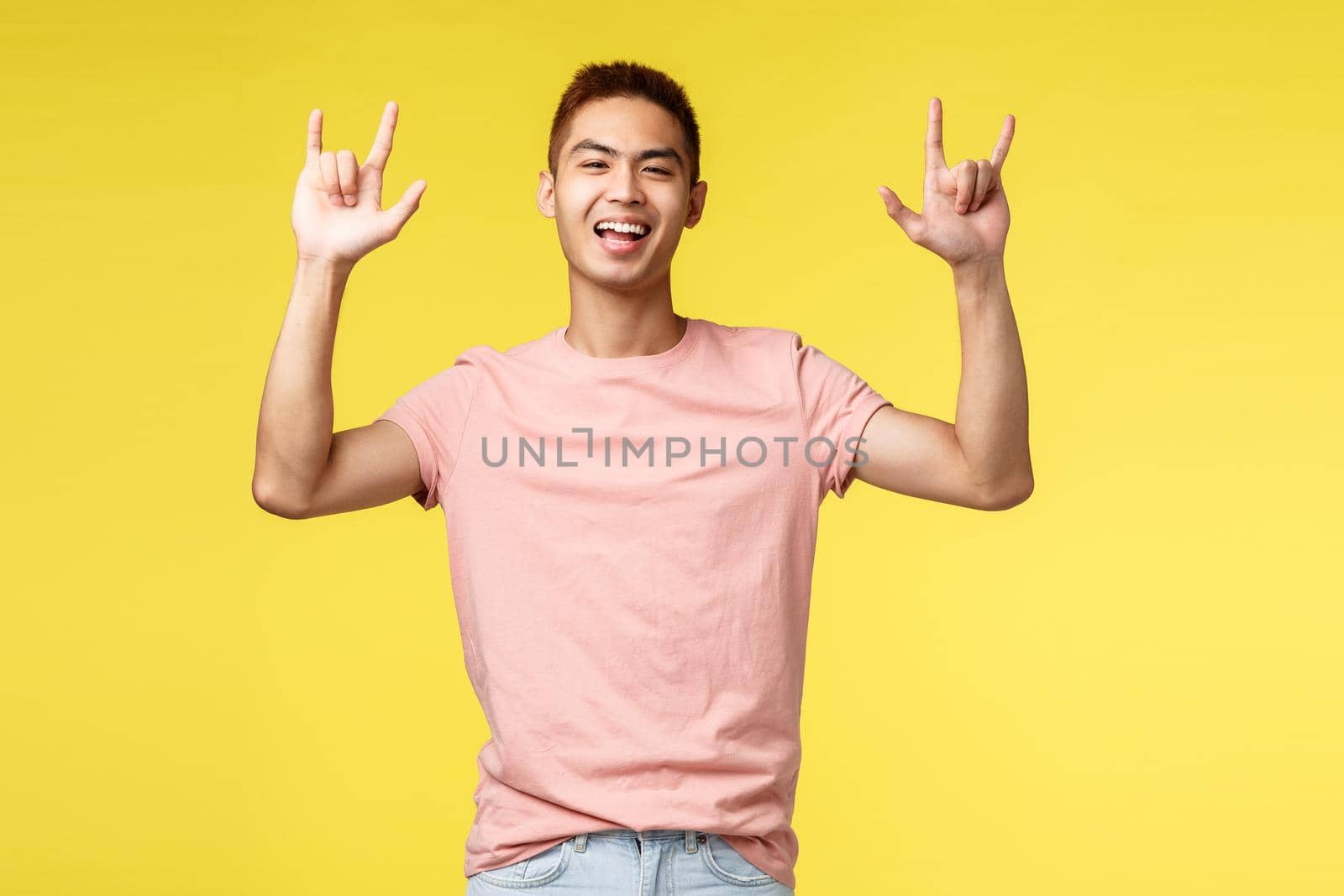 Lifestyle, education and people concept. Carefree handsome asian guy having fun, enjoying awesome summer campus party, show rock-n-roll sign and smiling broadly, partying, yellow background by Benzoix