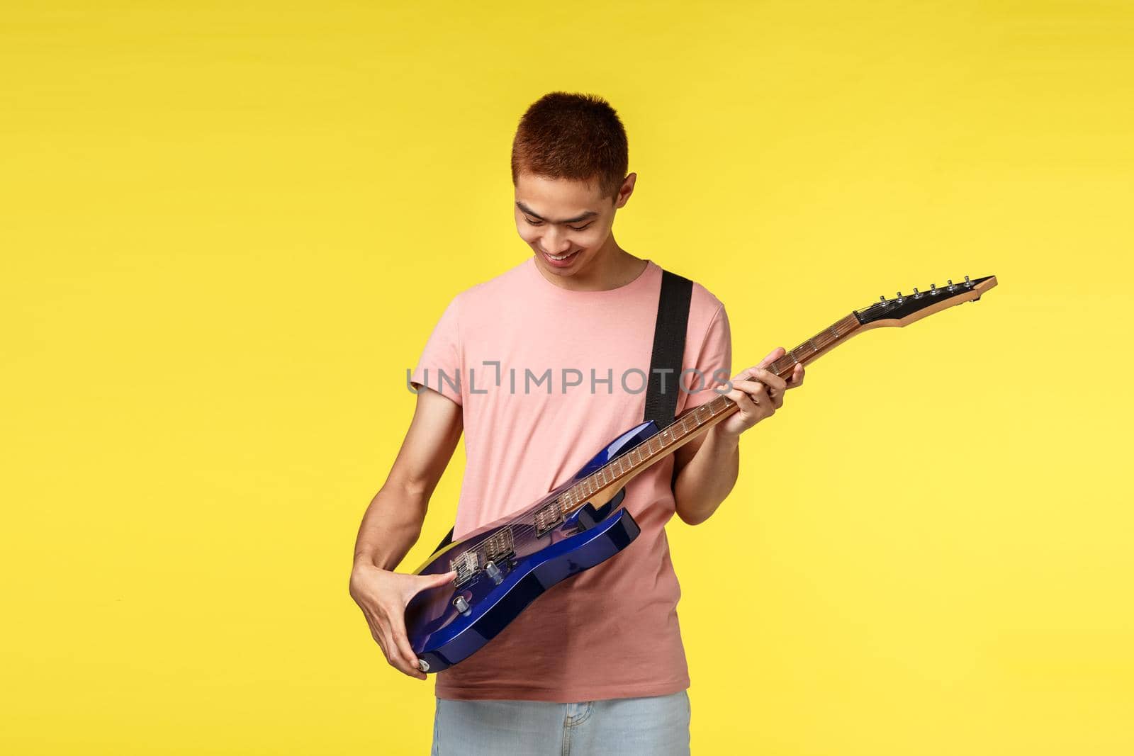 Lifestyle, leisure and youth concept. Portrait of enthusiastic asian guy receive new electric guitar as gift, smiling happy, look at intstrument satisfied and delighted, yellow background.