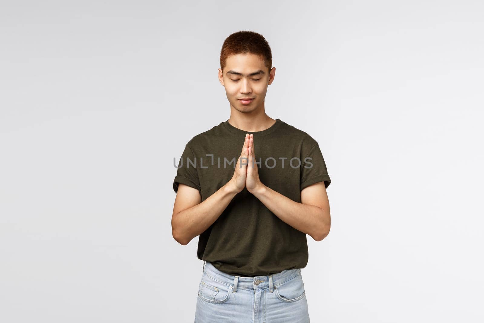 Portrait of hopeful young asian guy having faith, close eyes and smiling, clasp hands together in pray, pleading to god, want dream come true, supplicating, standing grey background.