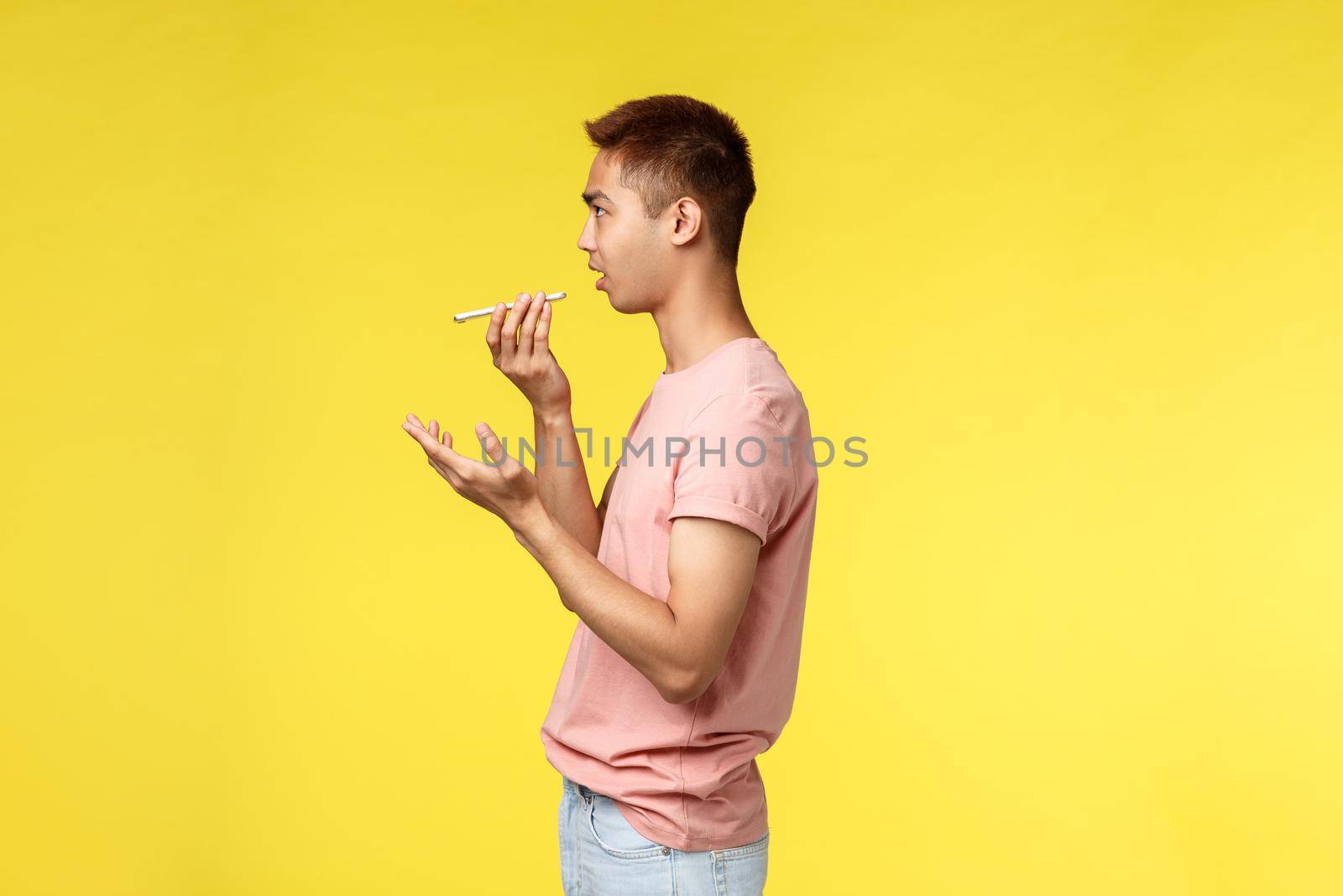 Technology, communication and lifestyle concept. Profile portrait of asian student guy leaving voice message using speaker, hold mobile phone close to mouth, gesturing, stand yellow background.