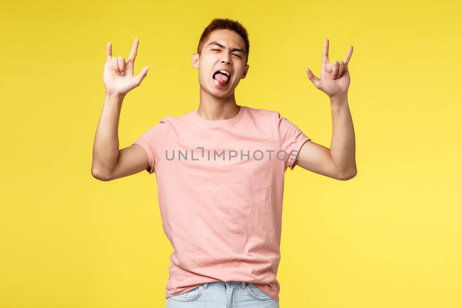 Lifestyle, education and people concept. Enthusiastic handsome and cool asian man in pink t-shirt, show tongue and rock-n-roll sign, having fun, partying, standing yellow background by Benzoix