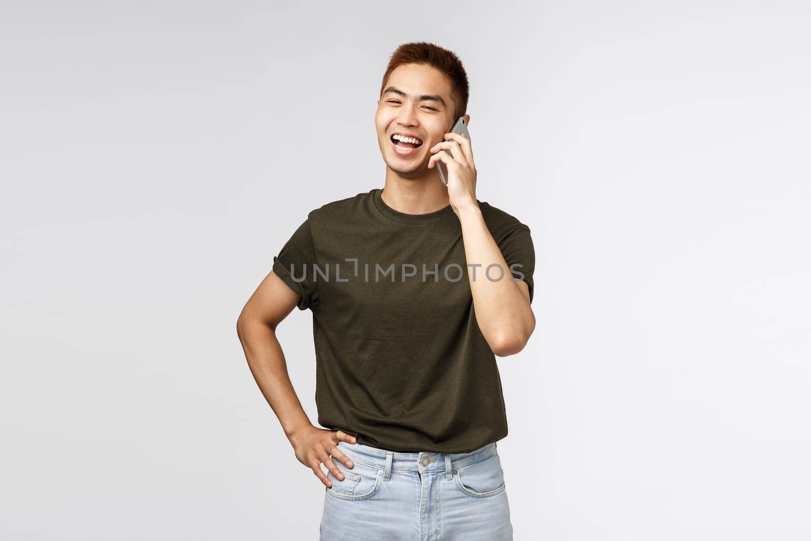 Technology, online lifestyle and communication concept. Portrait of cheerful laughing asian guy talking to friend via mobile phone, chuckle and smiling as looking away casually, grey background.