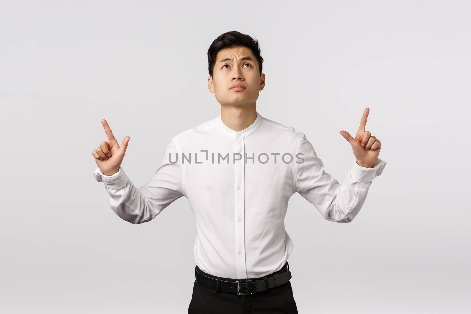 Skeptical asian businessman dont like what he see, standing hesitant and perplexed, frowning, sulking bothered, looking pointing up at terrible result, posing unsatisfied over white background.