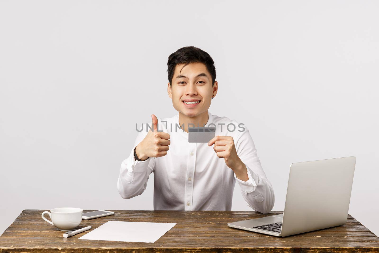 Satisfied good-looking asian office worker, manager sitting desk with documents, laptop and cup coffee, holding credit card, show thumbs-up and smiling, recommend buy online and use bank service.