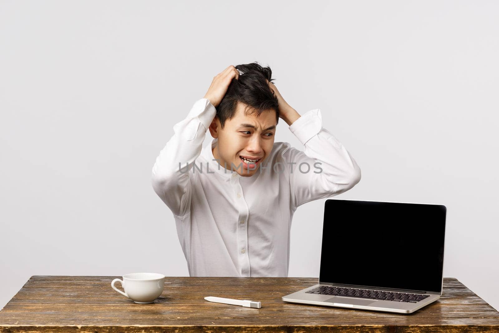 Alarmed and panicking chinese guy pulling hair from head sitting troubled and distressed, having huge problem, staring at laptop with anxious, nervous expression, sitting office white background.