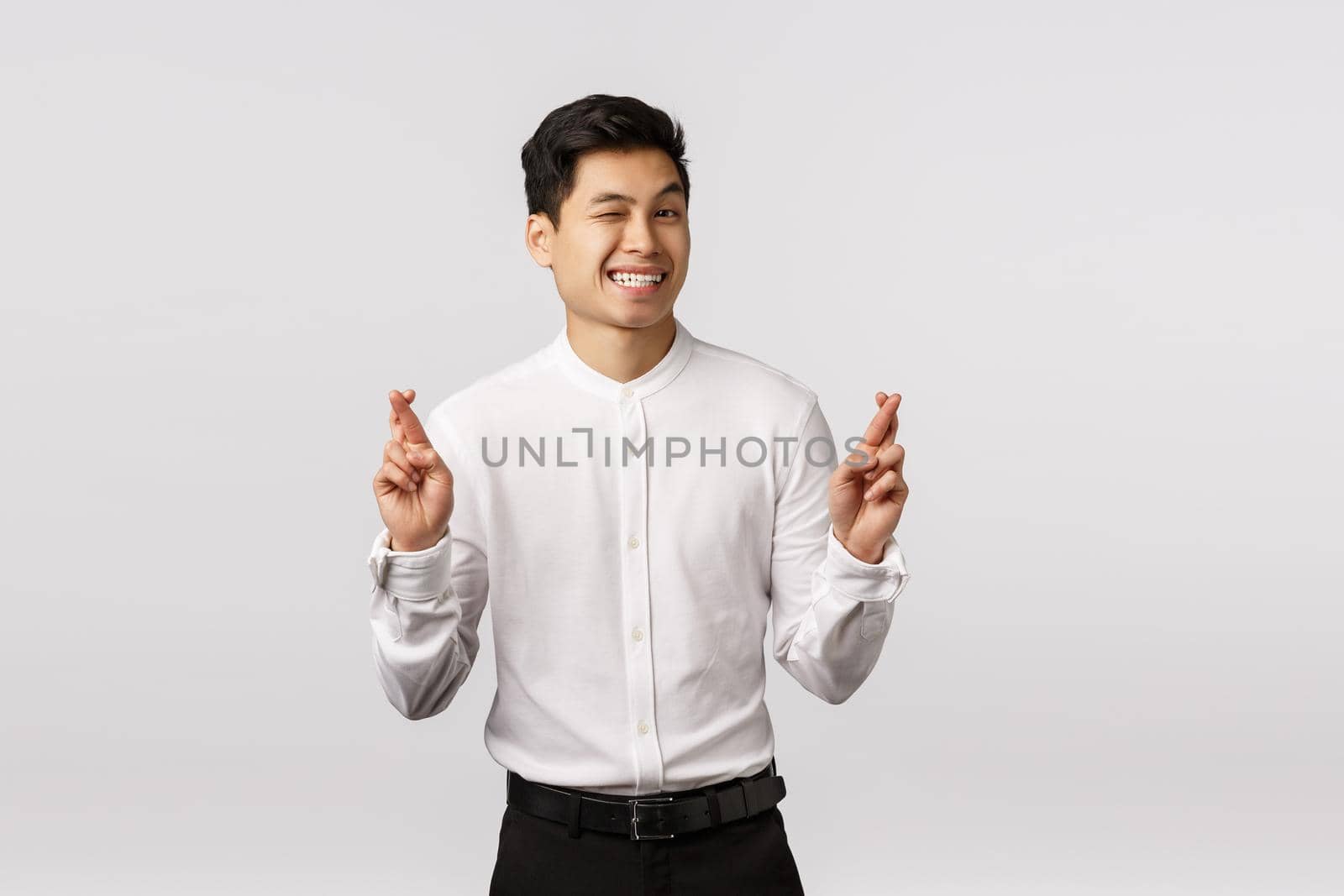 Cute hopeful optimistic asian male entrepreneur in white shirt, pants, wink camera cheeky, smiling assured everything be alright, cross fingers good luck, anticipate big deal at work signed.