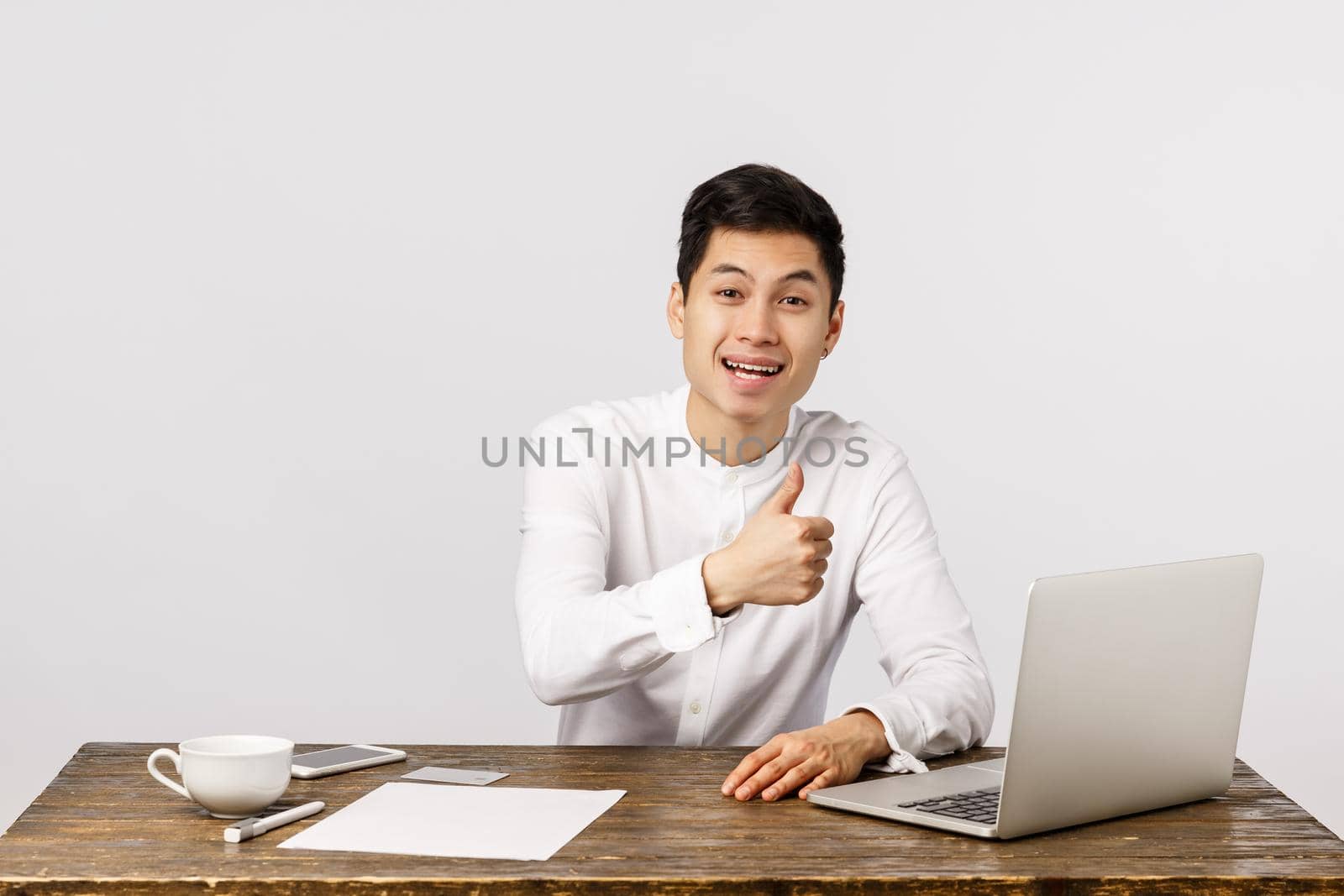 Finance, office and employer concept. Attractive cheerful asian young male entrepreneur in white shirt, sitting desk, drink coffee, approve something good, working laptop, documents, smiling pleased.