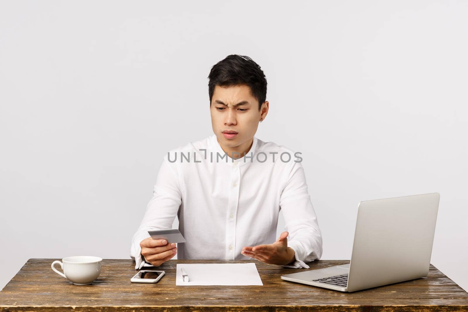 Distressed questioned and frustrated young asian guy sitting office at work, with documents and laptop, looking at credit card and complaining strange transactions, standing white background.