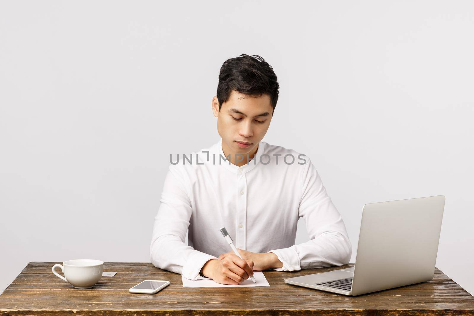 Business, work and interview concept. Busy serious handsome, young asian man sitting desk in office drink coffee, writing report, studying documents, use laptop, smartphone, white background.