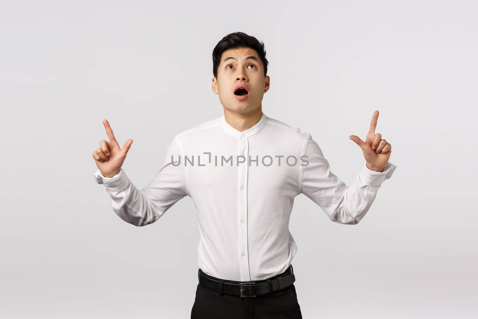Shocked and scared worried asian businessman see something falling down from sky, open mouth gasping shook and thrill, pointing looking up, stare speechless and amazed, standing white background.