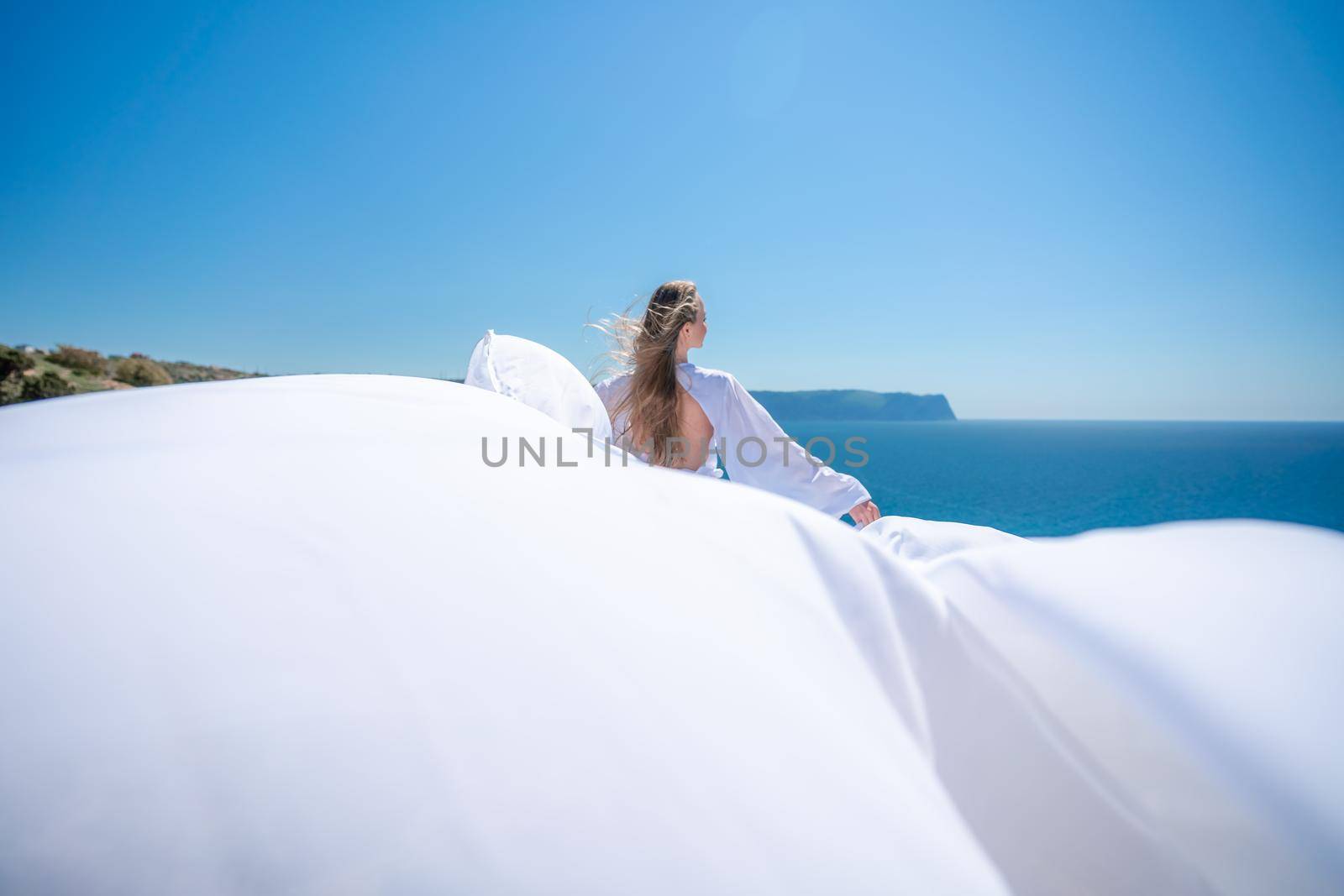 Blonde with long hair on a sunny seashore in a white flowing dress, rear view, silk fabric waving in the wind. Against the backdrop of the blue sky and mountains on the seashore