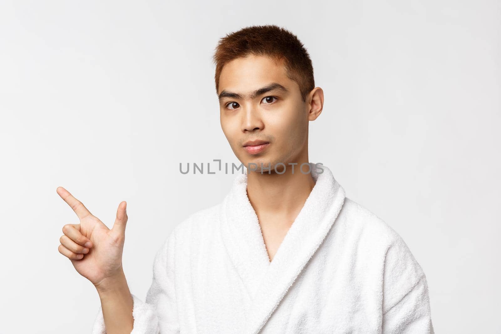 Beauty, spa and leisure concept. Portrait of young asian man inviting friends to hot springs, pointing fingers left and looking camera, show advertisement, wearing bathrobe, white background.