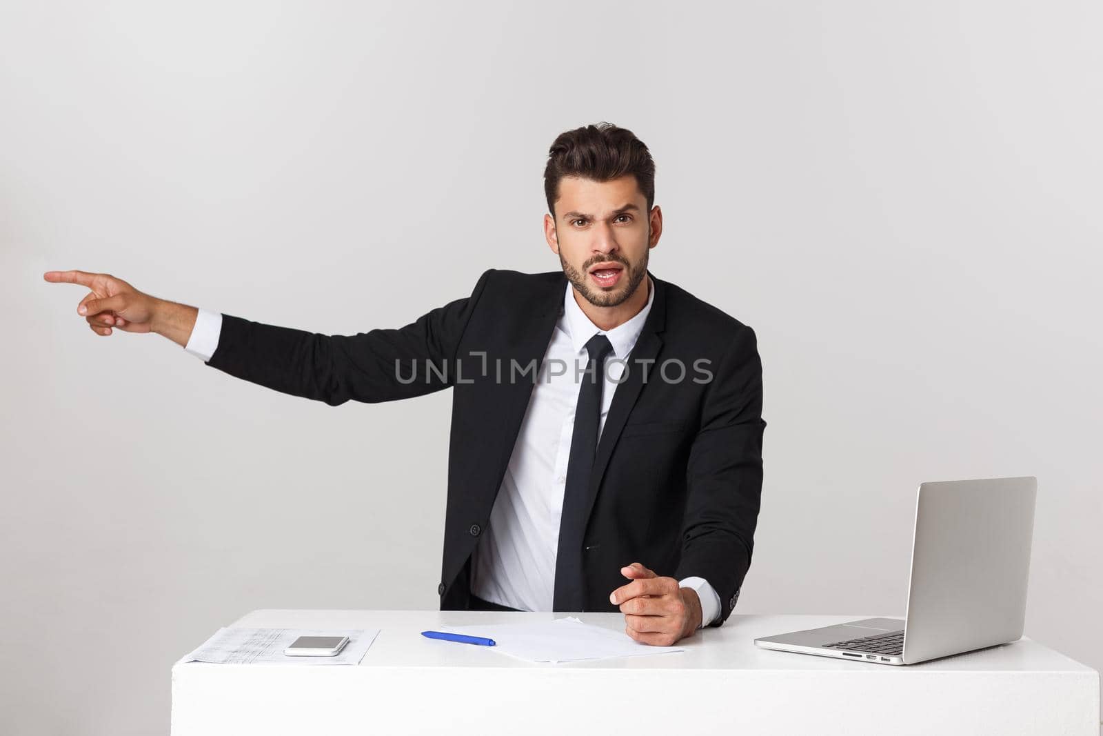 Angry businessman sitting at the table and screaming over gray background. Looking at camera.